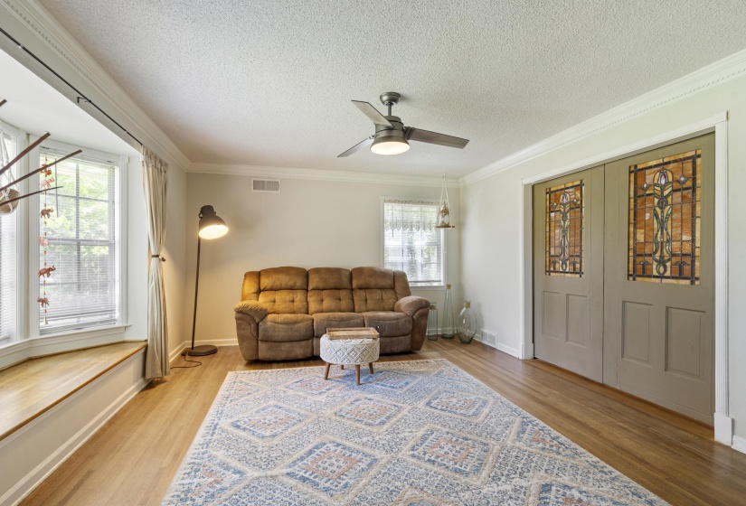 Living area featuring wood-finish flooring, a bay window with a built-in wood window seat, crown molding, a ceiling fan, and double doors with decorative stained glass panels
