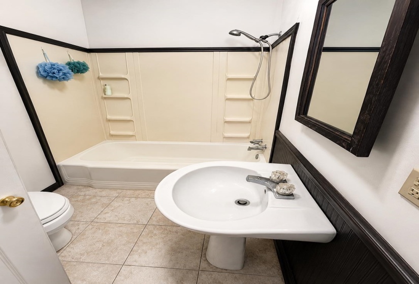 Bathroom featuring a white pedestal sink with a chrome faucet, a white toilet, and a bathtub with an integrated shower and cream-colored surround