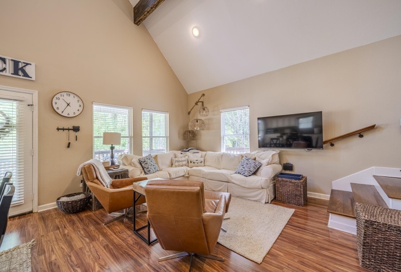 Vaulted ceiling living area featuring wood-finish flooring, multiple windows providing natural illumination, and a prominent wood beam accent