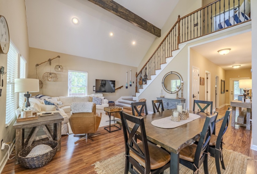 Open-concept living space featuring wood-finish flooring, a vaulted ceiling with exposed beam, and a staircase with an open balustrade leading to an upper level