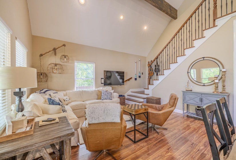 Bright living area featuring wood-finish flooring, a vaulted ceiling with recessed lighting, and a staircase with a dark wood handrail and black iron balusters