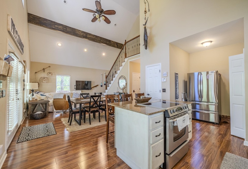 Open-concept kitchen and living area featuring high vaulted ceilings with exposed wood beams, wood-finish flooring, a kitchen island with a built-in range and granite countertop, and stainless steel appliances