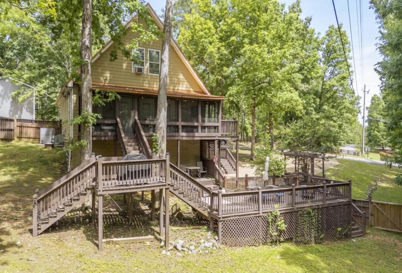 Multi-level outdoor decking and stairs frame this elevated home featuring a screened-in porch, a lower-level pergola, and mature surrounding trees
