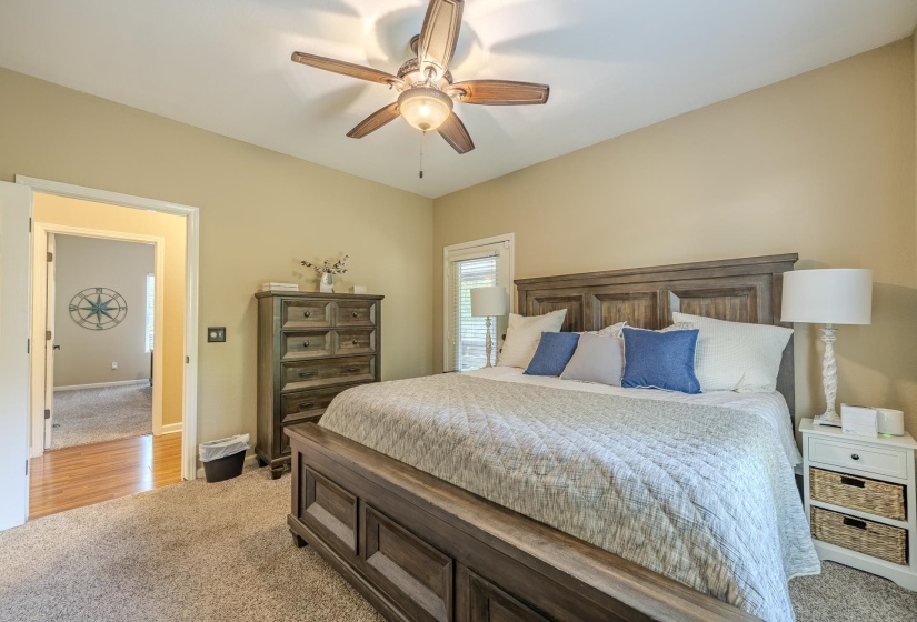 Spacious bedroom featuring neutral carpeting, a large window with natural light, a ceiling fan with light fixture, and an open doorway revealing wood-finish flooring in the adjoining space