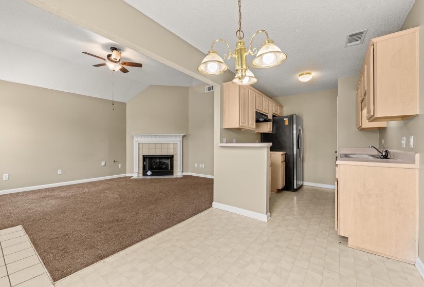 Combined living space featuring a kitchen with light wood-finish cabinetry and a living room with vaulted ceilings, a ceiling fan, and a tiled fireplace