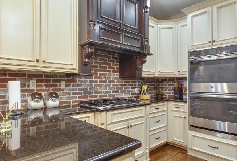 Kitchen featuring cream-toned cabinetry, dark wood-finish range hood, brick-look backsplash, dark countertops, and stainless steel double ovens