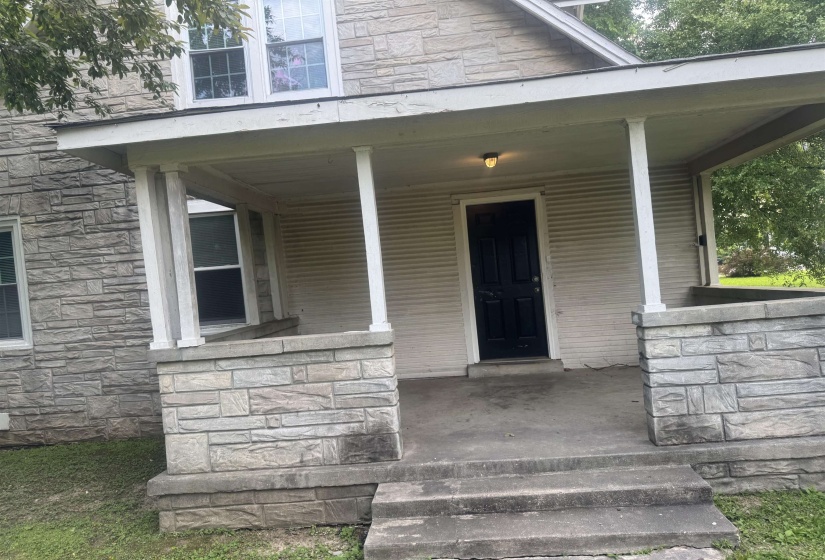 Covered porch entry featuring a dark paneled door, stone facade, column supports, and concrete steps