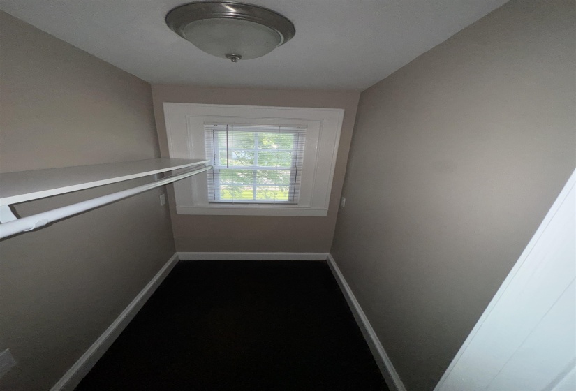 Compact closet space featuring a single window with blinds, dark flooring, and a built-in shelf with a hanging rod
