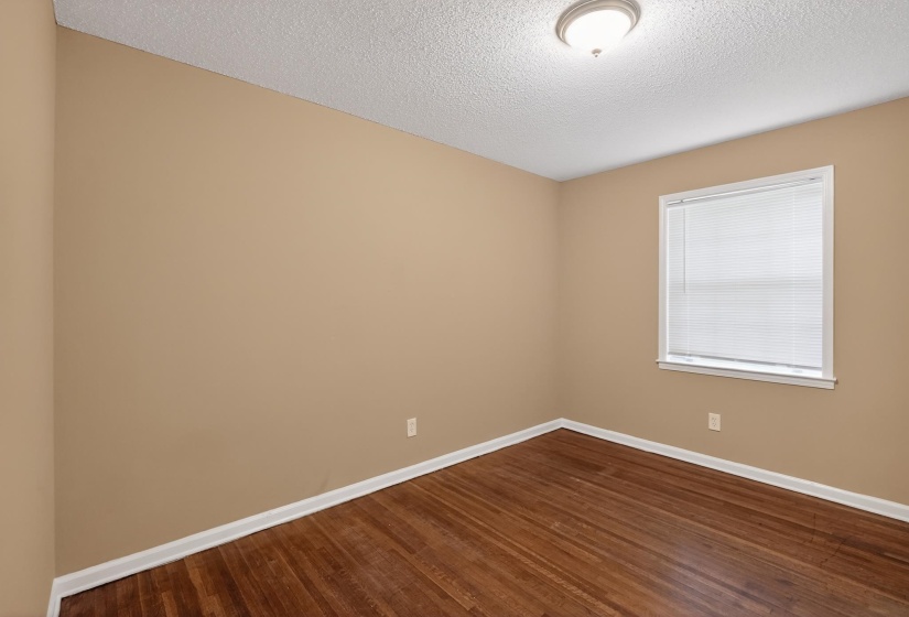Hardwood flooring throughout this room, featuring a window with blinds, white trim, and a ceiling-mounted light fixture