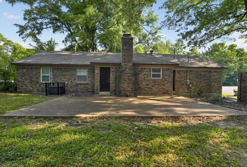 Exterior brick residence featuring a large concrete patio, a central brick chimney, a dark entry door, and multiple windows