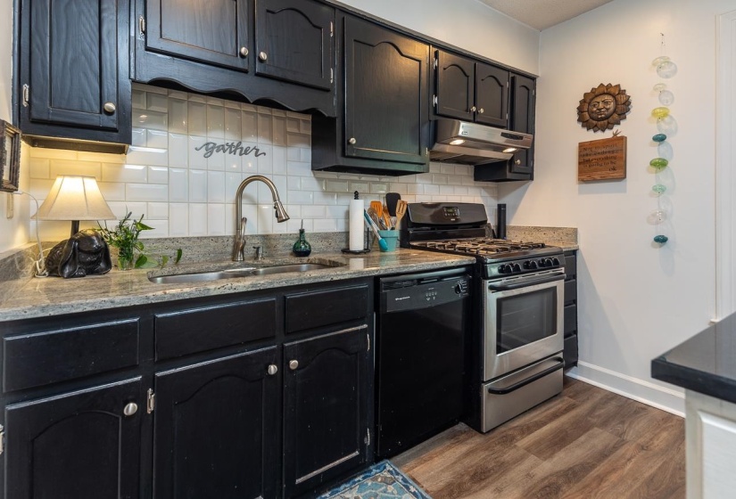 Kitchen with stainless steel gas range oven, dark wood-style flooring, under cabinet range hood, tasteful backsplash, and black dishwasher