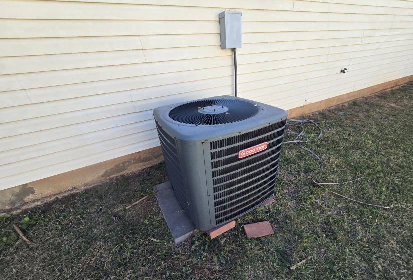 Goodman HVAC unit positioned on a concrete pad, against a cream-colored vinyl siding exterior