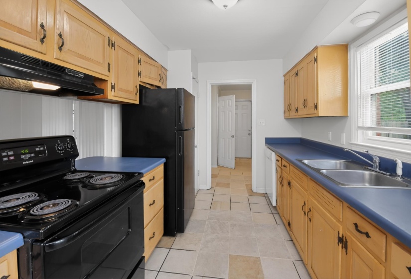 Galley kitchen featuring light wood cabinetry, blue countertops, a double basin stainless steel sink, black electric range, and a window with blinds