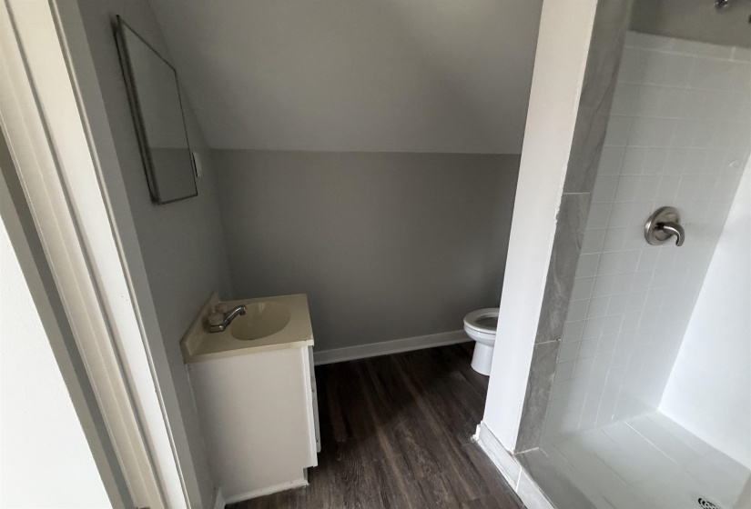 Compact bathroom featuring wood-finish flooring, a white vanity with an integrated sink, a rectangular framed mirror, and a white tiled shower with a brushed nickel fixture