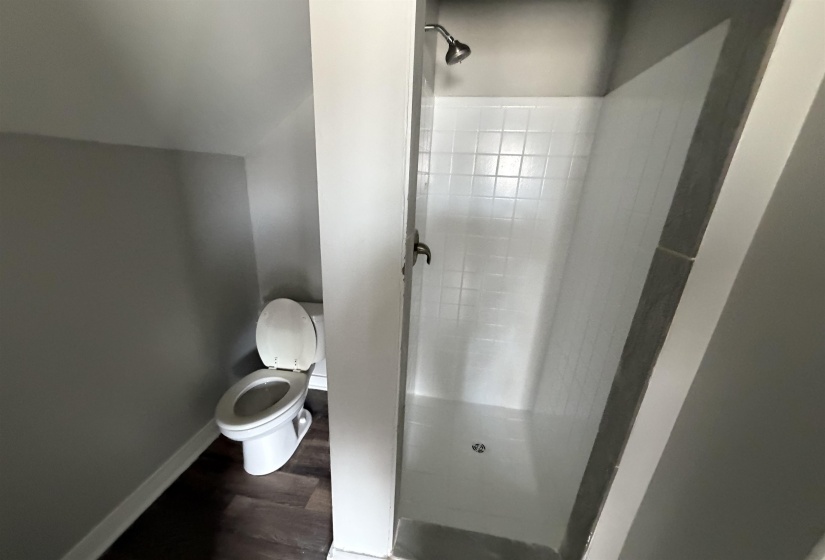 Compact full bathroom featuring a tiled shower with a chrome showerhead and control, a white toilet, gray wall paint, and dark wood-finish flooring