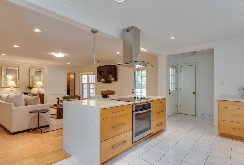 Modern kitchen island with integrated cooktop and oven, stainless steel range hood, light wood-finish cabinetry, and white countertop