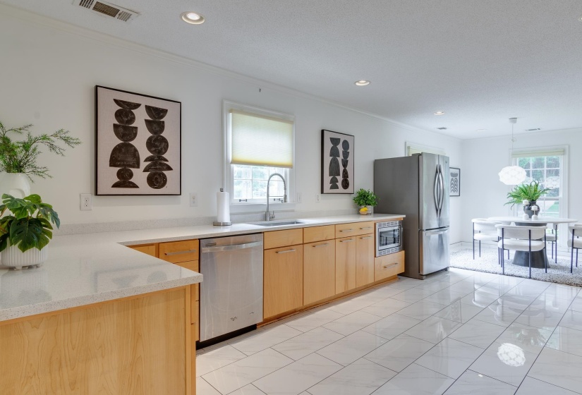 Open-concept kitchen featuring light wood cabinetry, stone countertops, stainless steel appliances, and white tile flooring