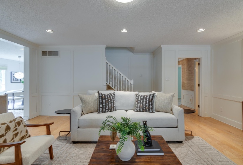 Recessed lighting illuminates the living area featuring wood-finish flooring, paneled wall detailing, and a staircase with a white railing