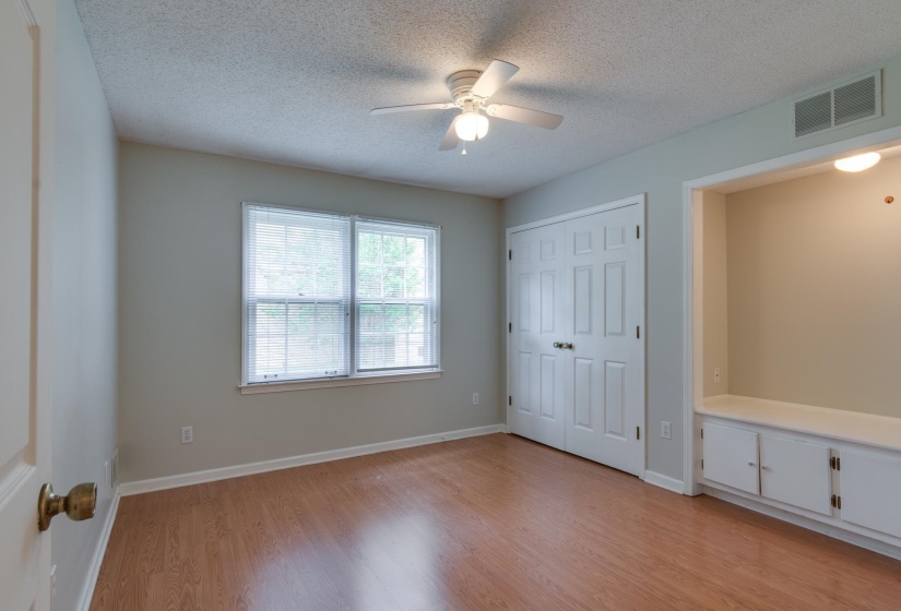 Interior room featuring wood-finish flooring, a double window with blinds, a ceiling fan with integrated lighting, a double-door closet, and a built-in storage bench with cabinets