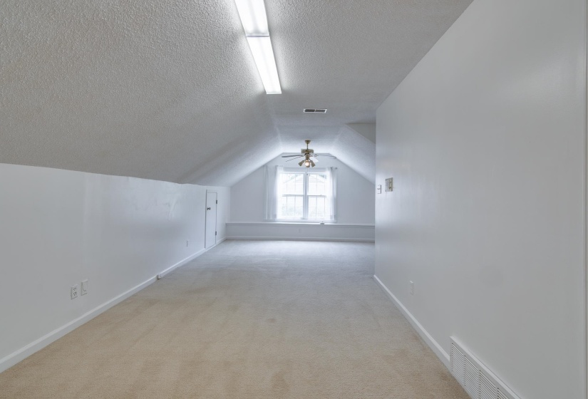 Expansive attic space featuring light beige carpeting, a window with natural light, a ceiling fan, and sloped ceilings