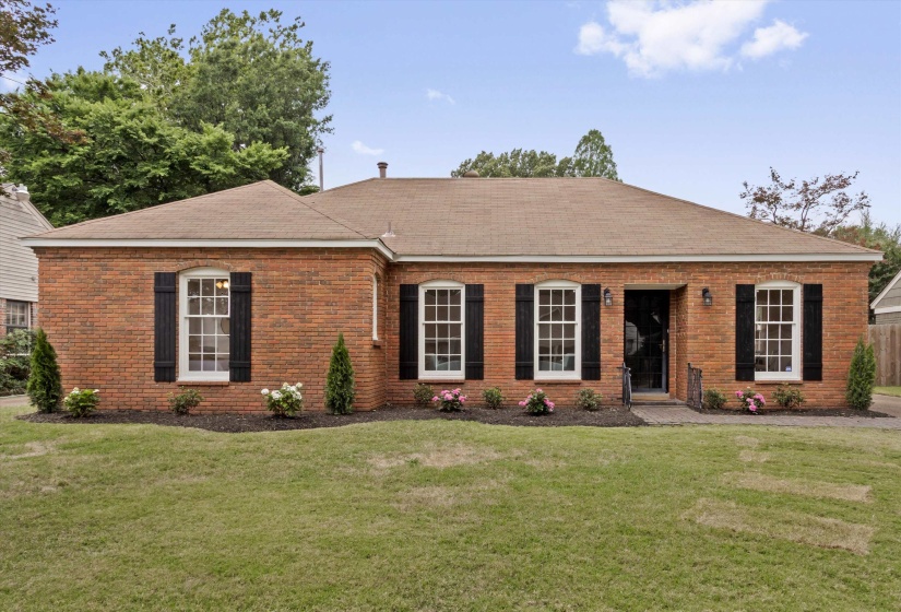 Brick exterior with a brown shingle roof, featuring white-framed windows with black shutters, a dark entry door, and a stone walkway