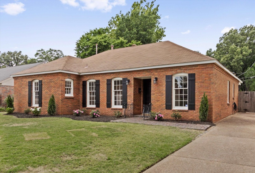 Brick exterior home featuring white-framed windows with black shutters, a brown shingle roof, and a concrete driveway