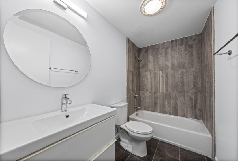 Contemporary bathroom featuring a white vanity with integrated sink, a large oval mirror, and a tub-shower combination with wood-look tile surround