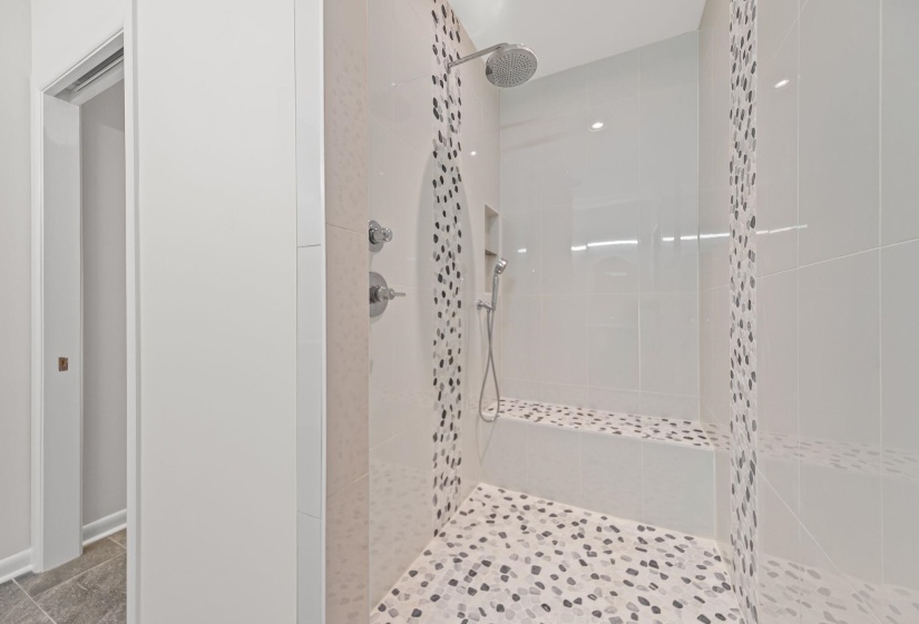 Walk-in shower featuring a rainfall showerhead, handheld sprayer, built-in bench, recessed shelf, and natural pebble tile flooring with coordinating accent stripes