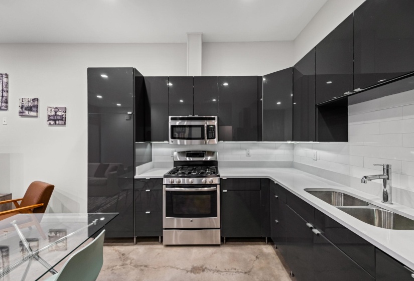 Modern kitchen featuring dark high-gloss cabinetry, stainless steel appliances, a subway tile backsplash, light countertops, and under-cabinet lighting