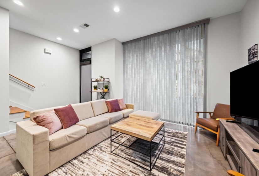 Spacious living area featuring high ceilings, recessed lighting, large vertical blinds, a visible stairway with wooden handrails, and a sleek wood-finish media console