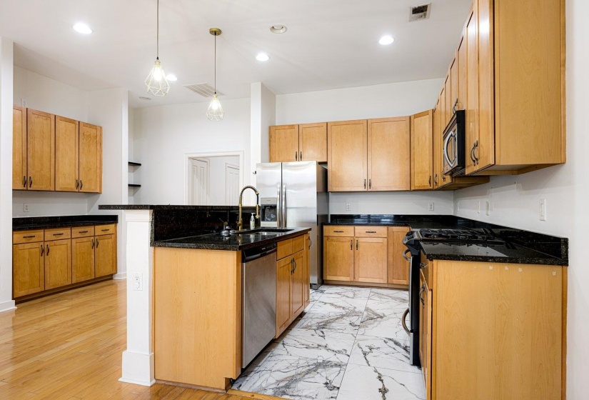 Kitchen with stainless steel appliances, dark stone countertops, decorative light fixtures, an island with sink, and wood finish cabinetry