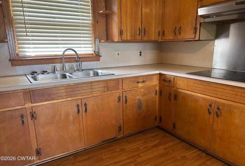 Kitchen featuring wood-finish cabinetry, a double basin stainless steel sink with a gooseneck faucet, light-toned countertops, and a built-in cooktop with an overhead stainless steel range hood