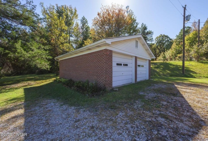 Detached brick two-car garage featuring two white garage doors with window inserts, white vinyl siding on the gable, and a gravel driveway