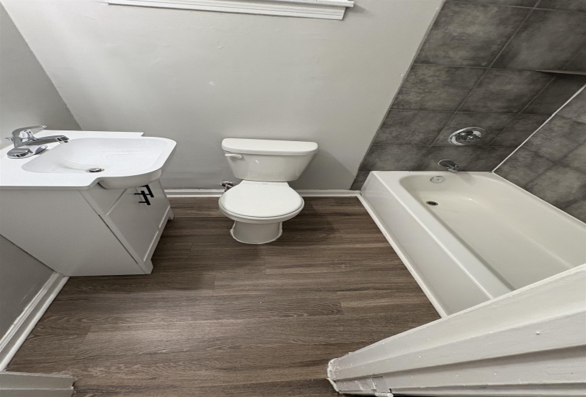 Bathroom featuring wood-finish flooring, a white vanity with a chrome faucet, a white toilet, and a white bathtub with dark gray wall tile surround