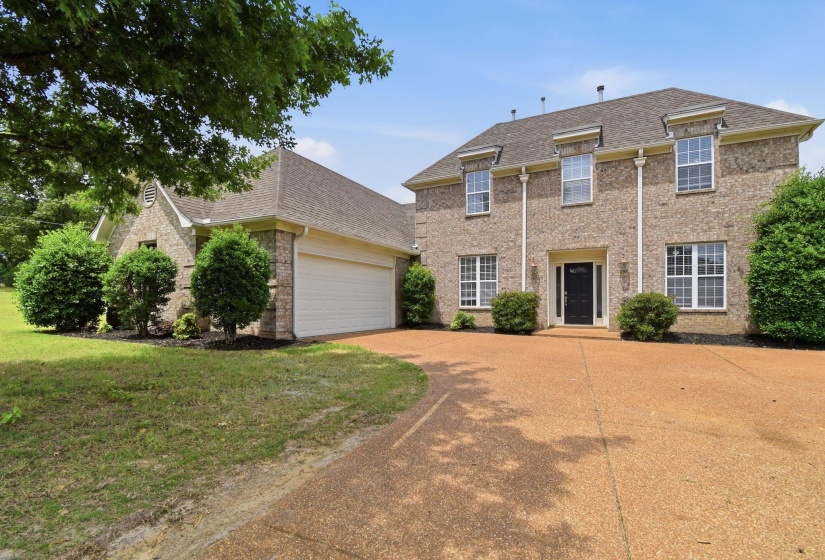 Traditional brick exterior featuring a multi-car garage, mature landscaping, a prominent front entry with a dark door, and a textured driveway