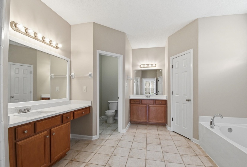 Spacious bathroom featuring twin vanity areas with wood cabinetry and white countertops, a large soaking tub, and neutral-toned tiled flooring