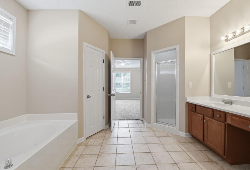 Spacious bathroom featuring neutral tile flooring, a large soaking tub, a separate glass-enclosed shower, a double vanity with a white countertop, and warm wood cabinetry