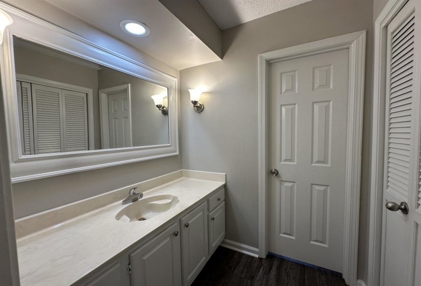 Bathroom vanity with a white countertop and integrated sink, framed mirror, wall-mounted sconces, recessed lighting, and wood-finish flooring
