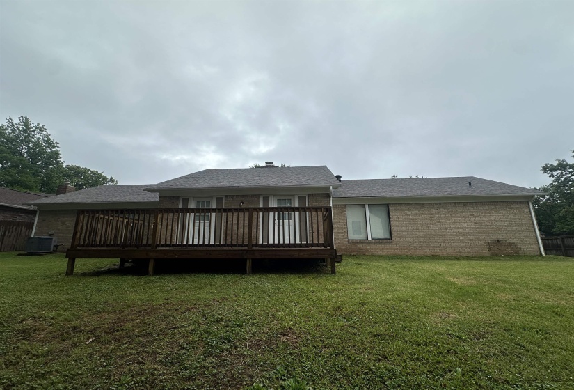 Expansive rear lawn featuring a wood-decked patio with balusters, light brick exterior, and a dark shingle roof