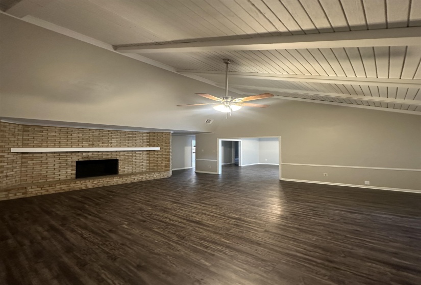 Vaulted ceiling with exposed beams, wood-finish flooring, brick fireplace with mantel, ceiling fan with integrated lighting, and neutral wall tones