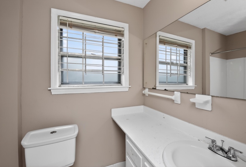 Bathroom featuring a white toilet, framed window with security bars, vanity with an integrated sink, wall-mounted mirror, and a shower/tub combination with white surround