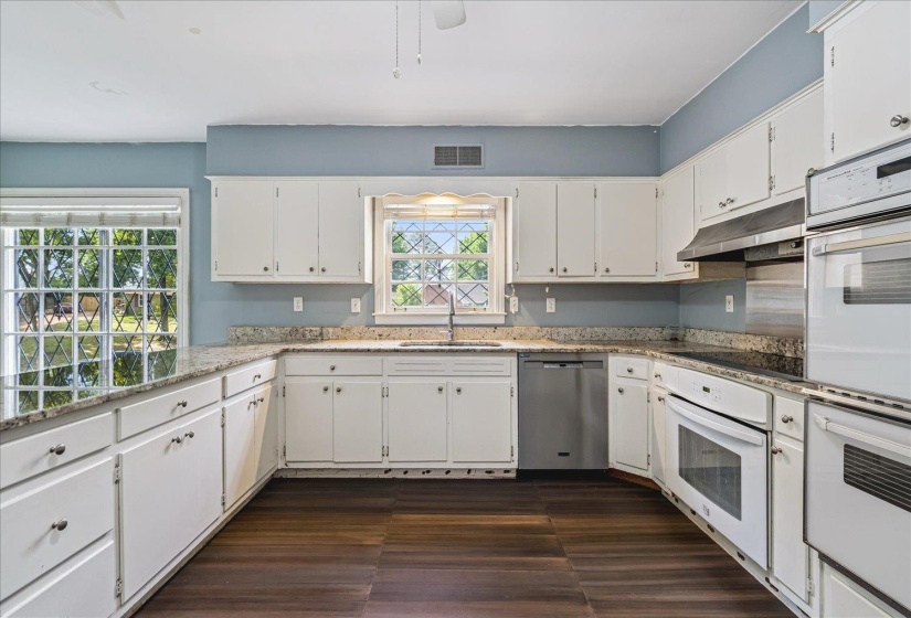Spacious kitchen featuring extensive white cabinetry, granite-finish countertops with matching backsplash, wood-finish flooring, a built-in oven, and a stainless steel dishwasher