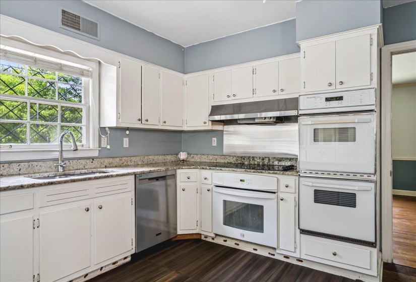 Well-appointed kitchen featuring white cabinetry, light-toned countertops, stainless steel dishwasher, integrated wall ovens, and wood-finish flooring
