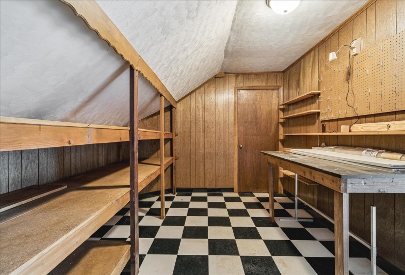 Attic storage room featuring black and white checkerboard flooring, wood-paneled walls, built-in shelving, and a built-in workbench with pegboard backing