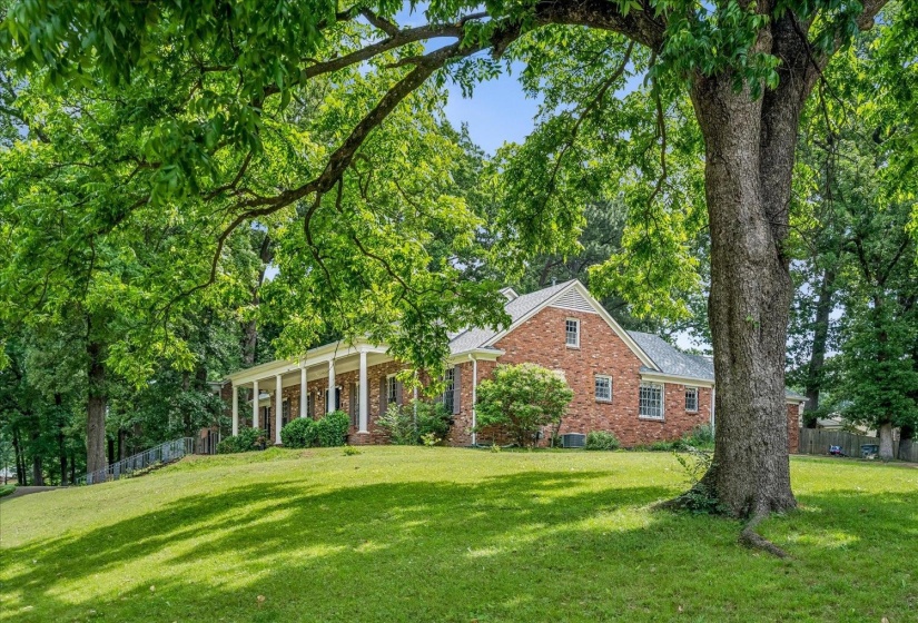 Brick exterior residence featuring a covered porch with white columns, a pitched shingle roof, and mature trees providing natural shading