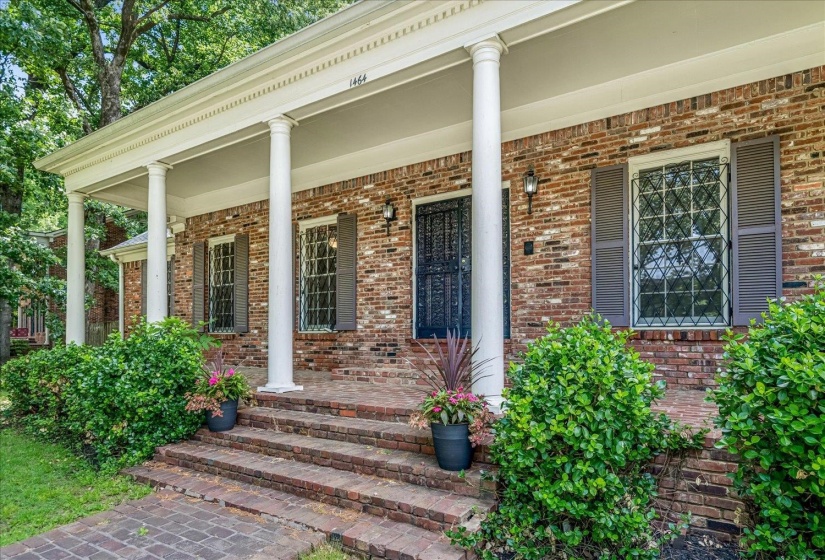 Brick exterior featuring a covered porch with white columns, a dark security screen door, and windows with decorative grilles and louvered shutters