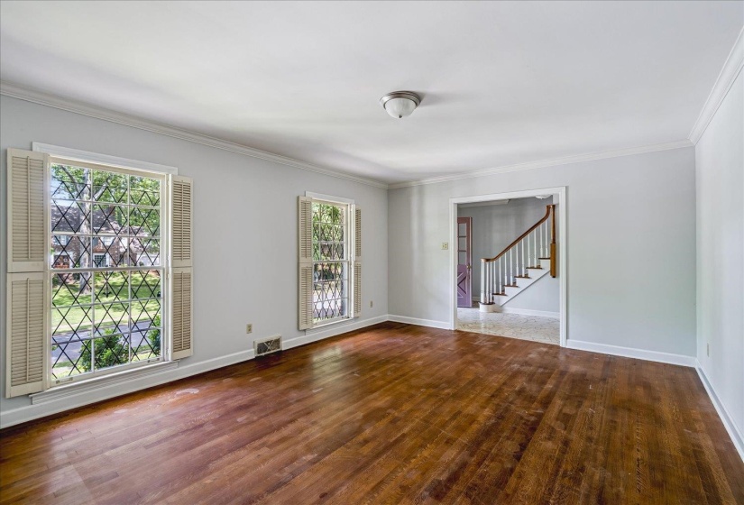 Spacious room featuring rich wood-finish flooring, two large windows with decorative security grilles and interior shutters, crisp white crown molding, and a warm neutral wall color
