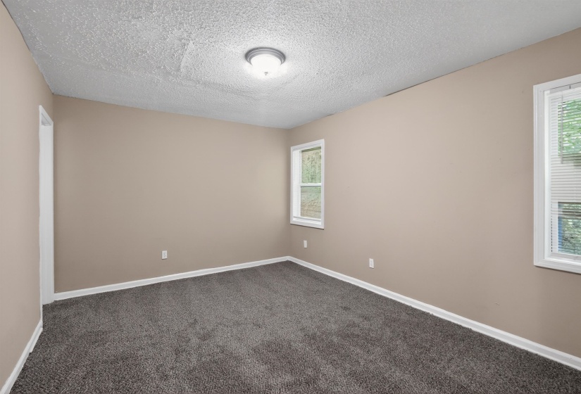 Spacious room featuring neutral-toned walls, textured ceiling, two windows with blinds, and dark gray carpeting