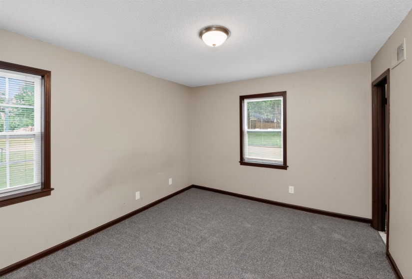 Neutral wall paint, two windows with dark wood trim and blinds, gray carpet, dark wood baseboards, and a ceiling-mounted light fixture