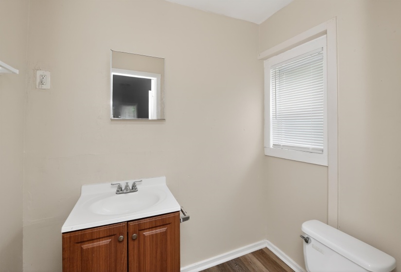 Bathroom featuring wood-finish flooring, a rectangular window with blinds, a white vanity with a single basin sink, and a wall-mounted mirror